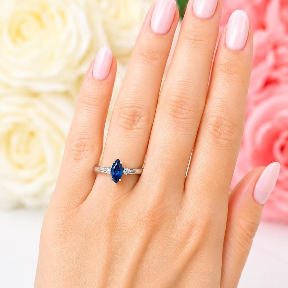 A woman's hand wearing a sterling silver 3-stone ring featuring a 10x5mm marquise cut lab blue sapphire flanked by round czs. Model image for size and styling reference — not a photo of the actual ring.