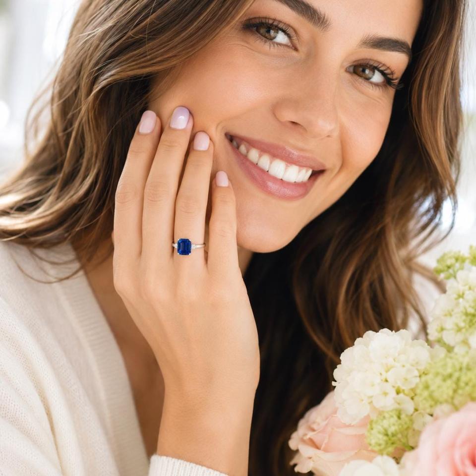 A woman wearing a sterling silver double prong solitaire ring featuring an emerald cut lab blue sapphire flanked by a round brilliant cz on each side. Model image for size and styling reference — not a photo of the actual ring.